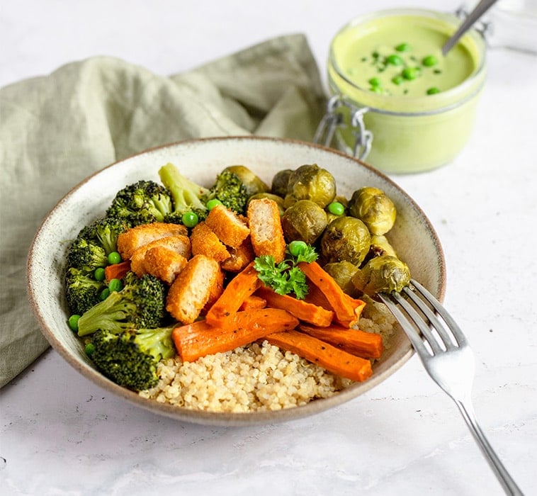 A bowl with pieces of coated fish, Brussels sprouts, carrots, couscous, broccoli and a pea dressing